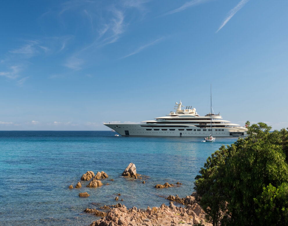 This stunning wide-angle view captures the beautiful vessels on the water at the luxurious Costa Smeralda resort in Sardinia, Italy. The expansive view showcases a massive super-yacht on the right, contrasted by the smaller vessels nearby, highlighting the sheer size and grandeur of the larger boat. The breathtaking scenery of the Mediterranean is visible in the background, with the sun-drenched hills and lush vegetation framing the tranquil turquoise and blue waters of the bay. This image perfectly captures the summertime paradise and exclusive luxury destination that Costa Smeralda is known for. Visitors to this awesome location can immerse themselves in the beauty of the Mediterranean, enjoy luxurious accommodations, and experience the ultimate in relaxation and serenity.