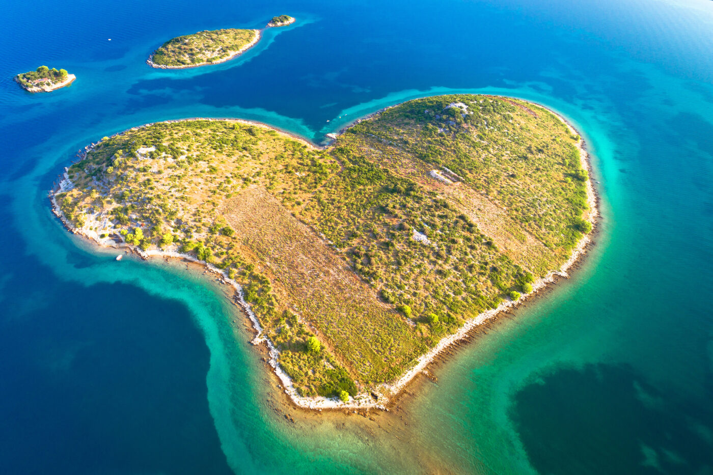 Aerial view of a heart-shaped island surrounded by turquoise and deep blue waters, with patches of green vegetation and rocky shoreline, creating a stunning natural formation.