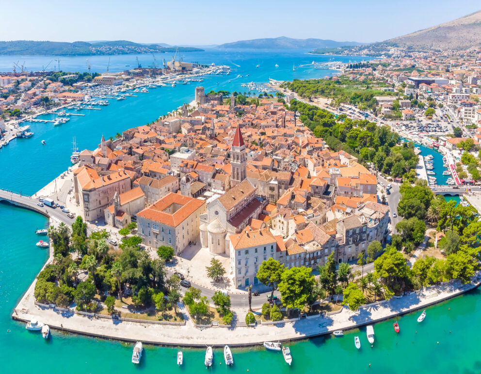 Aerial view of the historic town of Trogir, Croatia, featuring medieval stone buildings with orange rooftops surrounded by turquoise blue water and boats docked along the shore.