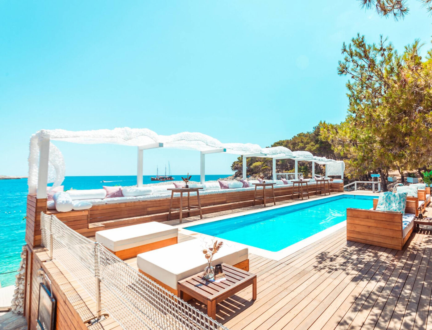 Luxury beach club with a wooden deck, pool, and white canopy daybeds overlooking the turquoise Adriatic Sea on a sunny day, surrounded by pine trees.