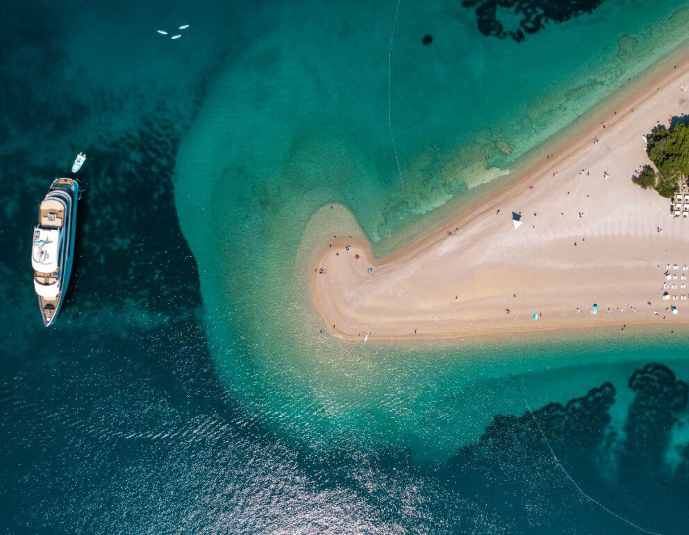 Aerial view of Zlatni Rat Beach (Golden Horn) in Croatia, showing its distinctive V-shaped sandbar stretching into turquoise waters, with a yacht anchored nearby and sunbathers along the shore.