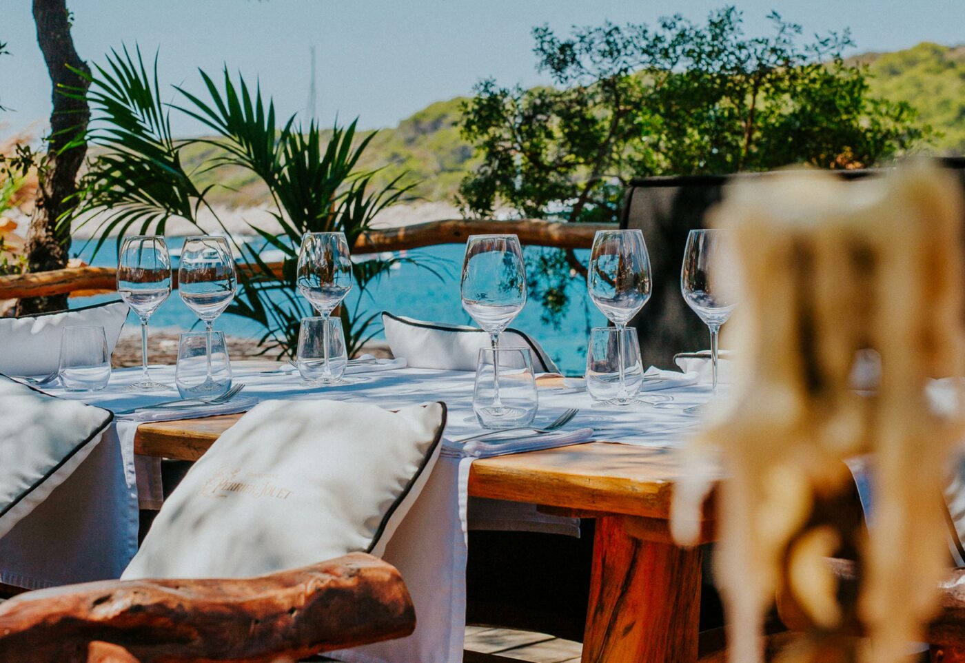 Elegant seaside restaurant table set with white linens, wine glasses, and rustic wooden chairs, overlooking turquoise water and surrounded by lush greenery.