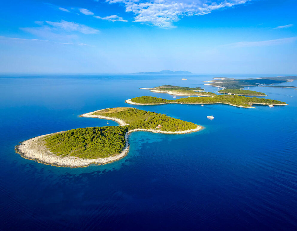 Aerial view of the Pakleni Islands in Croatia, featuring a chain of small green islands surrounded by calm, deep blue Adriatic waters under a clear sky.