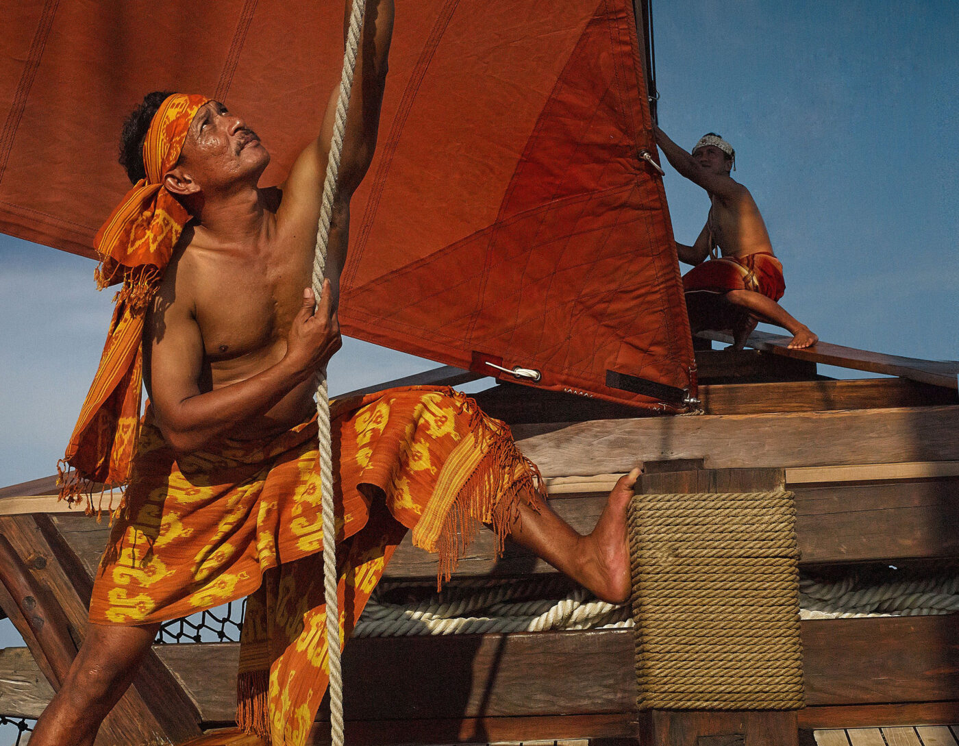 Traditional Indonesian sailors hoisting sails on a wooden phinisi boat.