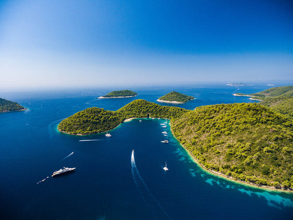 Aerial view of Lastovo, Croatia, showing lush green islands surrounded by deep blue Adriatic waters with yachts and boats sailing between them under a clear sky.