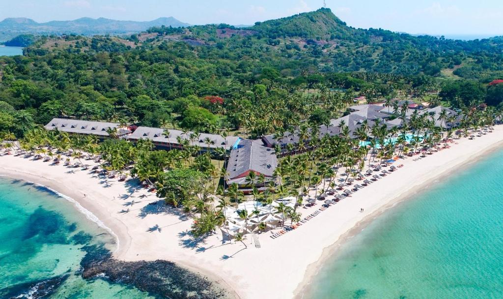 aerial view of the Andilana Beach Resort in Nosy Be, Madagascar. The resort is situated on a peninsula between two white sand beaches with turquoise water. The complex includes several buildings with dark roofs, surrounded by lush green vegetation and palm trees. There are sun loungers and umbrellas set up along the beach and next to a swimming pool