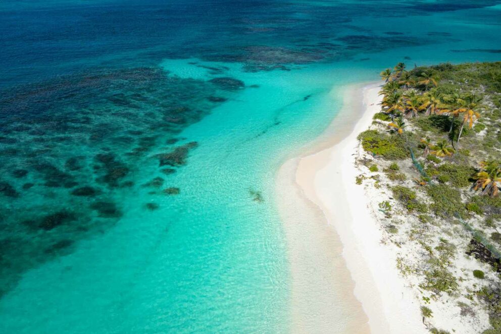 Aerial view of a white sand tropical beach next to clear turquoise ocean water and coral reefs.