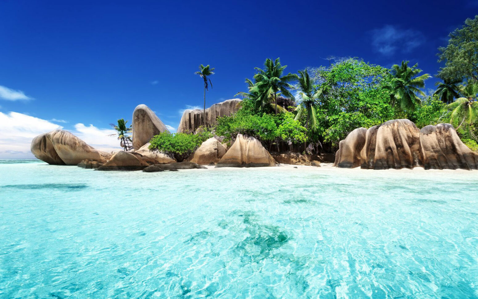 Stunning beach with crystal-clear turquoise water in the foreground, leading to a stretch of powdery white sand. large, smooth granite boulders that create a dramatic and picturesque landscape. Lush green vegetation and palm trees frame the beach.