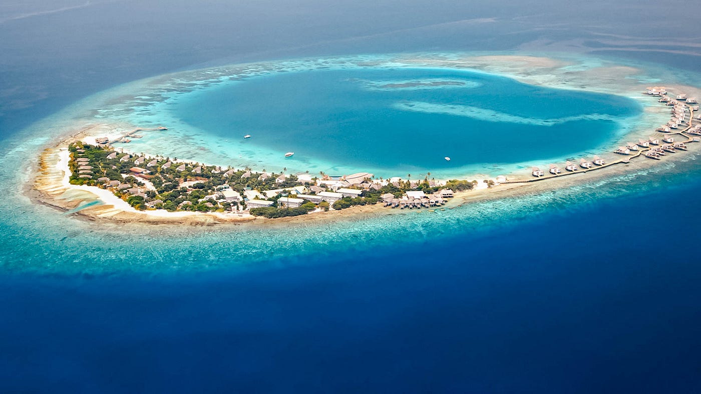 aerial photograph of a tropical island resort, in the Maldives. The island is situated within a large, circular coral reef, creating a shallow, turquoise-colored lagoon. On the island, there are numerous buildings, including bungalows and other resort structures, some of which extend out over the water. The island is covered in green vegetation and surrounded by a narrow strip of white sand. The reef separates the calm, light-blue lagoon from the deep, dark-blue ocean surrounding the atoll.