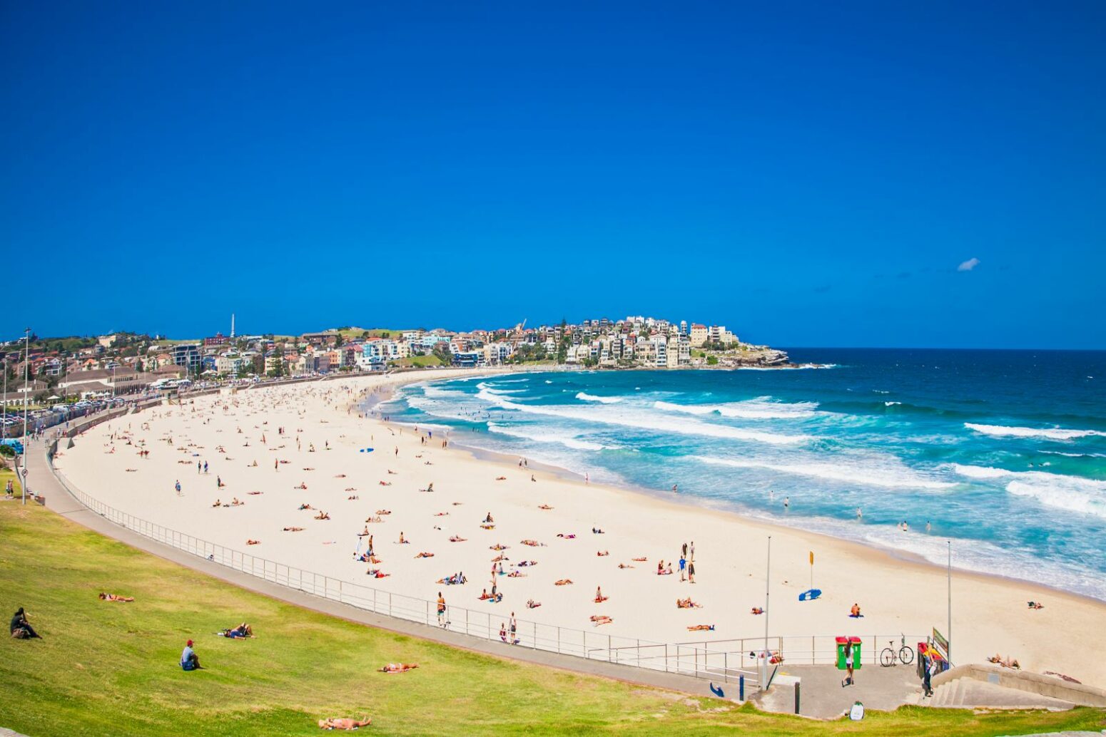 Sunny day at Bondi Beach, Sydney, with people sunbathing on the sand and swimming in the turquoise waves, surrounded by a coastal town under a clear blue sky.