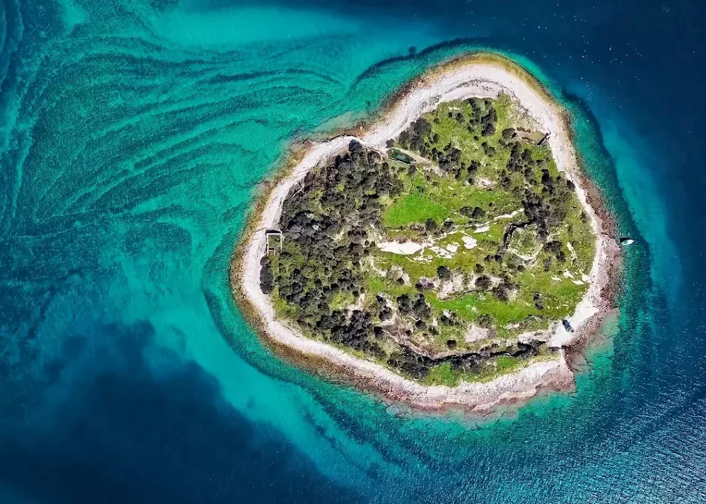 aerial view of a small, heart-shaped island in the middle of a turquoise and deep blue sea. The island is part of the Brijuni National Park in Istria, Croatia.