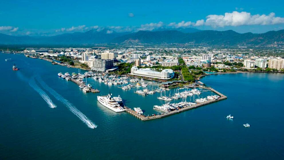 Aerial view of Cairns and Port Douglas in Australia, featuring a vibrant marina filled with yachts and boats, clear blue waters, and the cityscape stretching toward lush green mountains under a bright blue sky.