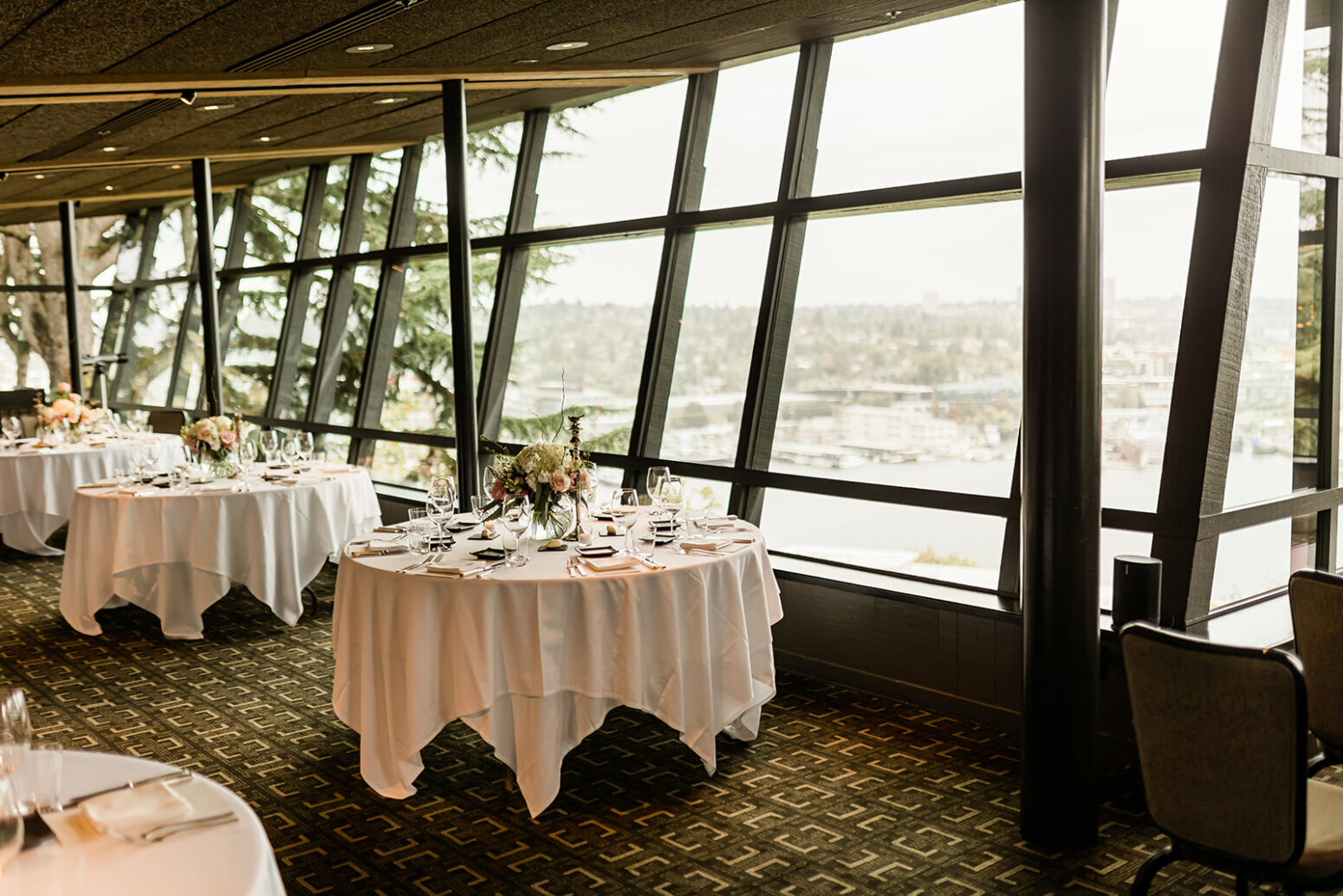 Elegant dining room at Canlis restaurant in Seattle, Washington, with round tables set with flowers and large windows overlooking the city view.