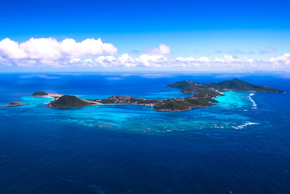 Aerial view of a lush green tropical island surrounded by turquoise and deep blue ocean water, with a visible coral reef and white clouds in a bright blue sky.
