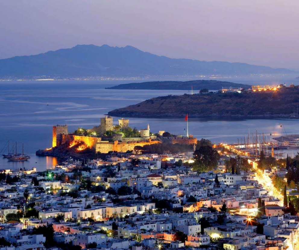 A nighttime photo of the city of Bodrum, Turkey, showing the medieval Castle of St. Peter on a peninsula, brightly lit. The city's white buildings line the coast, and a marina with yachts is visible to the right. A large body of water and a distant mountain range are in the background under a dark blue sky