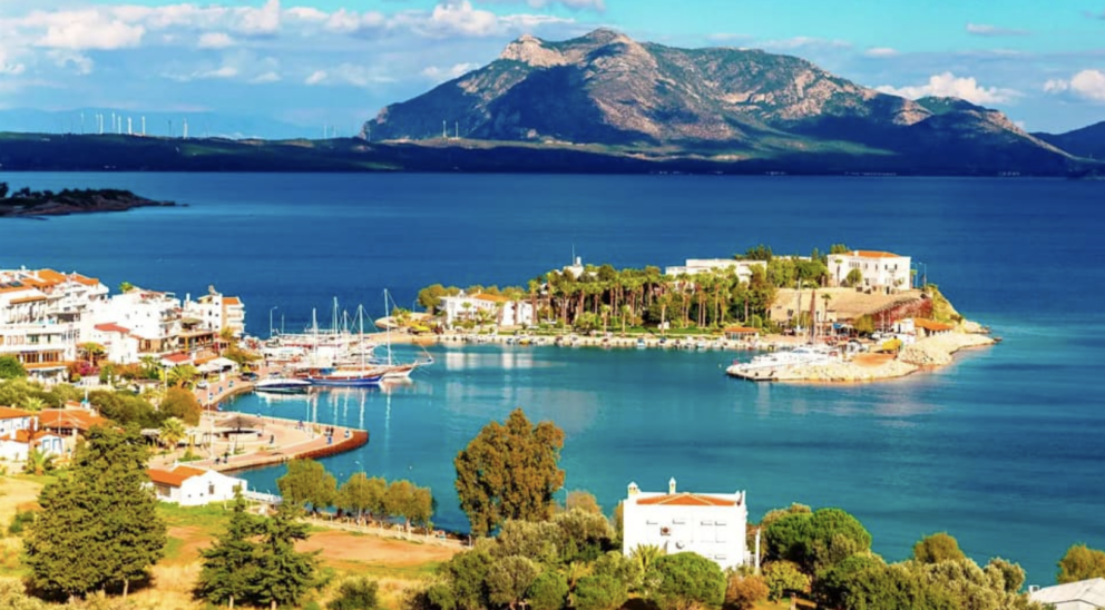 A scenic view of a coastal town with white buildings and a harbor full of boats, with a small island in the center of a bay. A large, green mountain is visible in the background under a blue sky with white clouds.