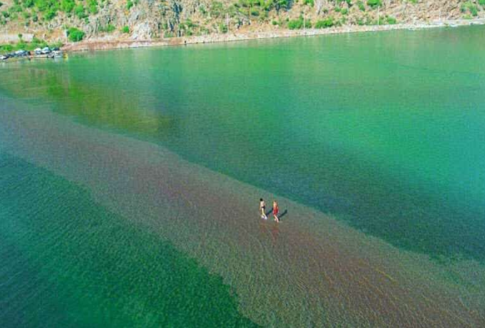Two people walk along a narrow sandbar in the middle of a shallow, green-blue body of water. The water is clear enough to see the sandy bottom. A green, rocky hillside is visible in the background. The scene is an aerial view.