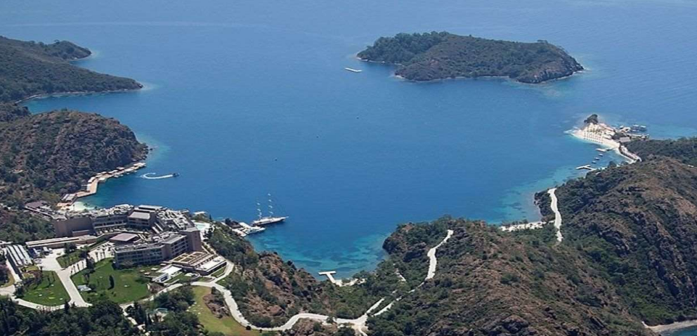 An aerial view of D-Hotel Maris resort on a bay with clear blue water. The resort buildings are nestled on the left side of the bay, surrounded by lush green hills. A yacht is docked near the hotel, and a winding road is visible on the hills. Small islands are located in the bay.