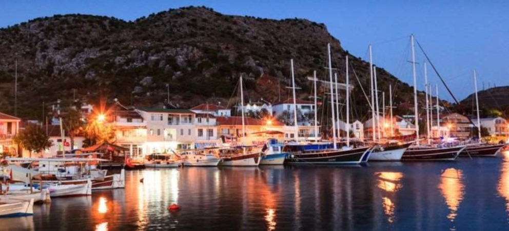 A nighttime photo of a harbor with several boats and yachts docked along a waterfront with buildings and a mountain in the background. The lights from the buildings and boats reflect on the water.