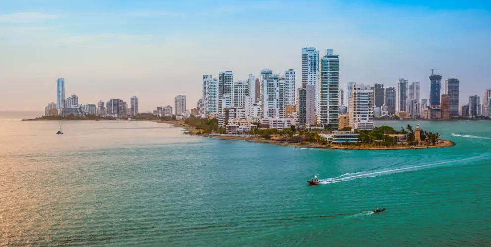 Panoramic view of Cartagena, Colombia, with modern skyscrapers along the Caribbean Sea and boats sailing on turquoise water at sunset.