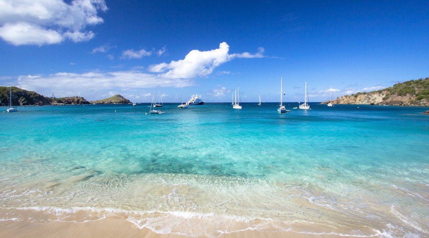 A beautiful tropical beach with clear turquoise water, a sandy shore, and several sailboats anchored in a bay surrounded by green hills under a blue sky with white clouds.
