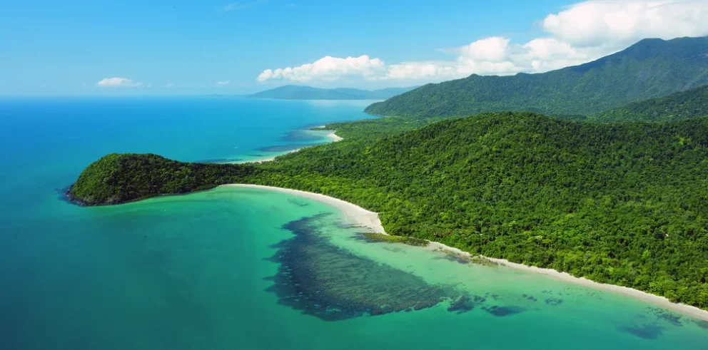 Aerial view of the Daintree Rainforest in Australia, where lush green forested hills meet turquoise ocean waters and white sandy beaches under a bright, partly cloudy sky.