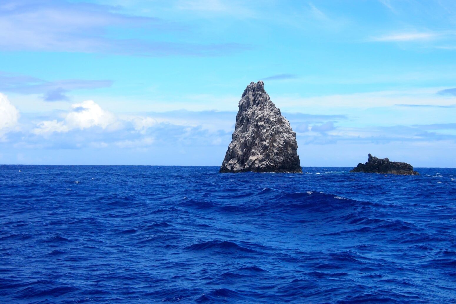 A large, jagged rock formation (Diamond Rock) in the middle of a deep blue ocean under a partly cloudy blue sky.