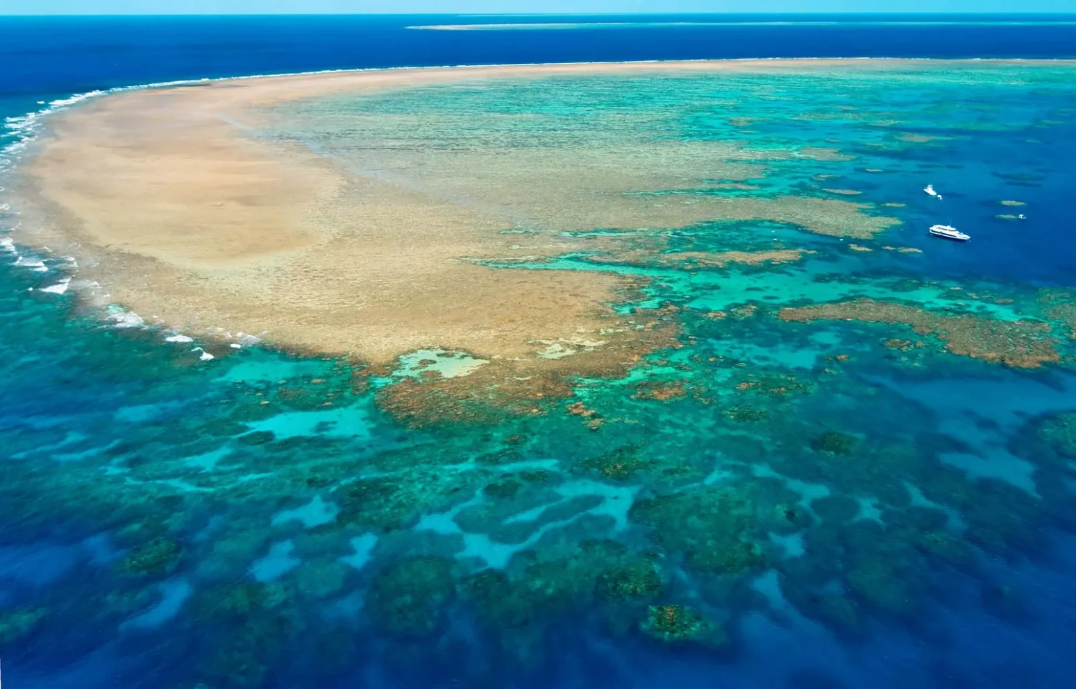 Aerial view of the Great Barrier Reef in Australia, showcasing turquoise blue waters, coral formations, and sandy reef patches, with a few boats floating nearby.