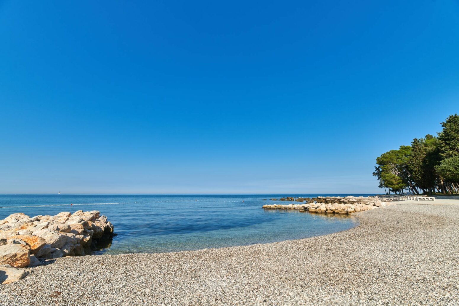 A serene, clear blue sea meets a rocky beach under a bright, cloudless sky. The calm water is bordered by large rocks on one side and a line of green trees on the other, creating a peaceful coastal scene.