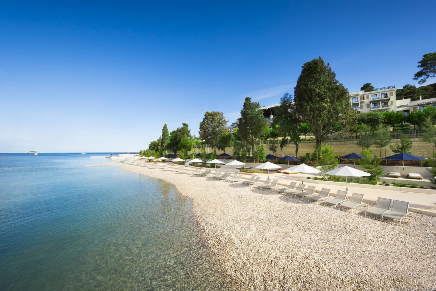A tranquil, pebbled beach with lounge chairs and umbrellas lines a calm, turquoise sea under a clear blue sky. In the background, a modern hotel is nestled on a lush, green hillside. The scene depicts Mulini Beach in Rovinj, Croatia.