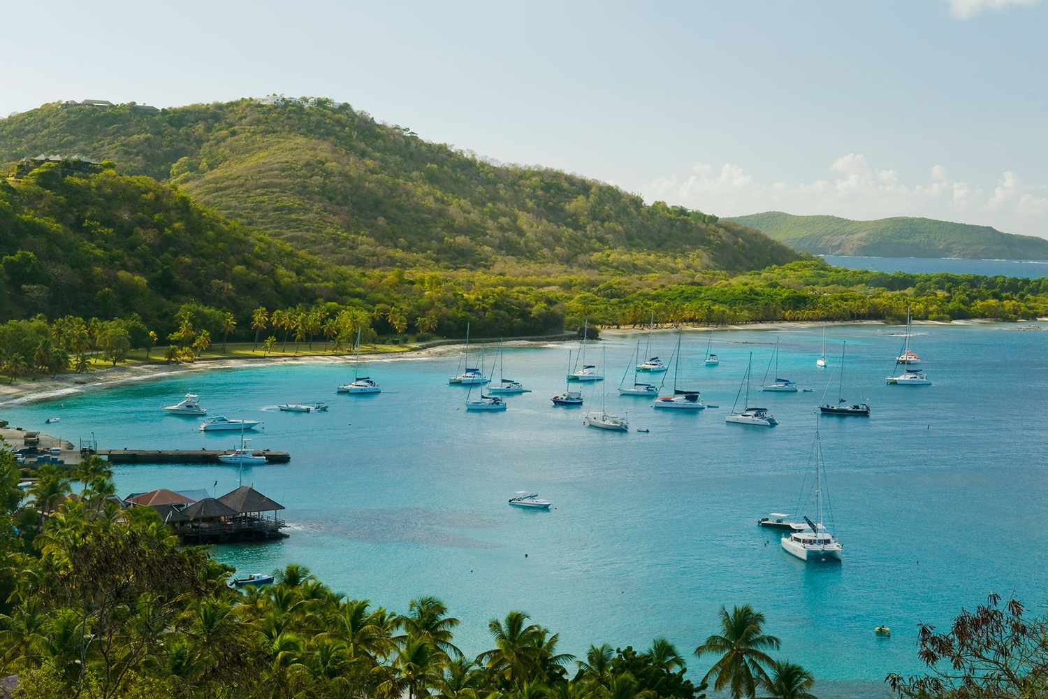 A scenic view of a tropical bay with turquoise water, anchored boats, a sandy beach, and lush green hills.