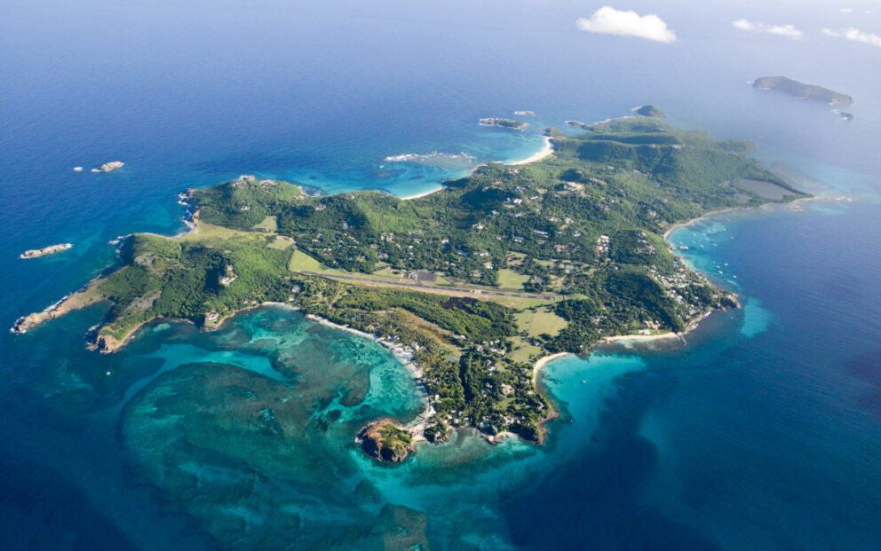 Aerial view of the lush, privately-owned island of Mustique in the Caribbean, featuring green hills, white sand beaches, and a central airstrip.