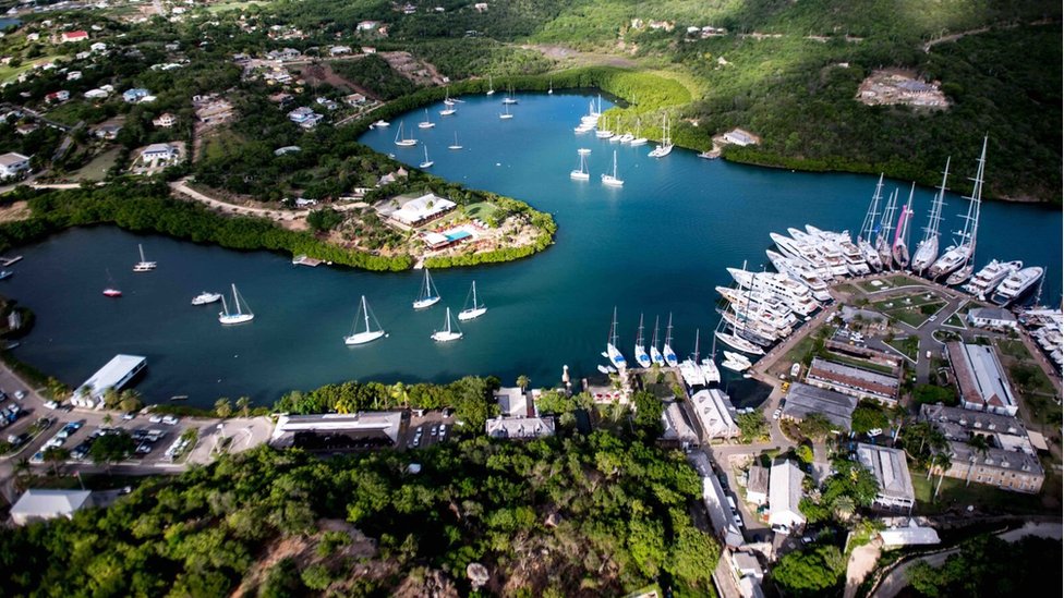 An aerial view of a picturesque, historic naval base and harbor, now a UNESCO World Heritage site, featuring numerous white sailboats and yachts docked in a calm, blue bay surrounded by lush green hills and colonial-style buildings.