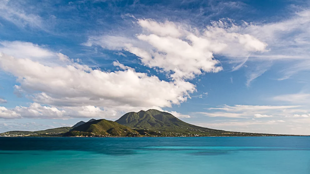 A lush green volcanic island, Nevis, rising from a turquoise blue Caribbean sea under a partly cloudy sky.