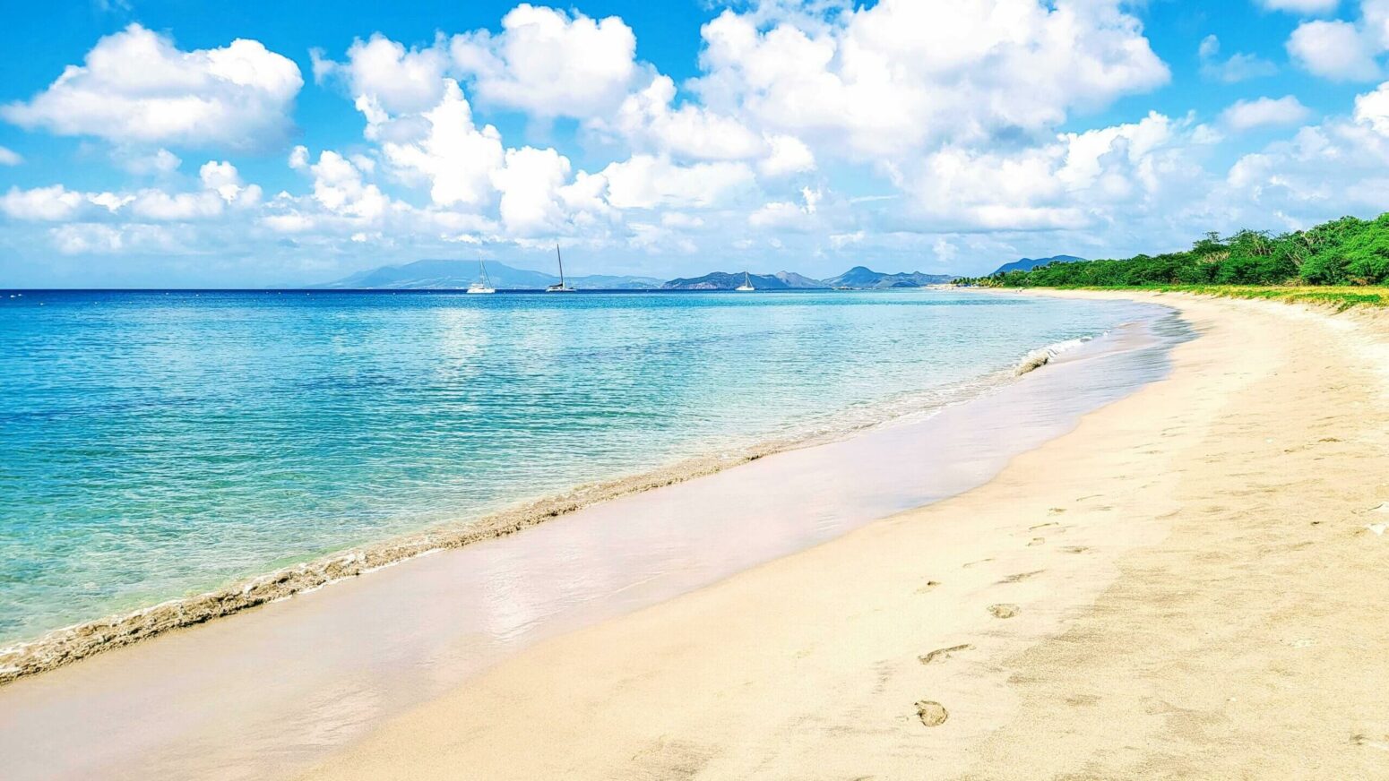 A wide, sandy beach with turquoise water, green trees on the right, distant islands, and a blue sky with white clouds.
