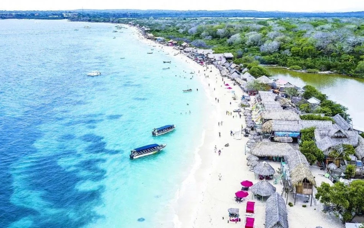 Aerial view of Playa Blanca in Cartagena, Colombia, with turquoise water, white sand, beach huts, boats, and people enjoying the tropical shoreline.