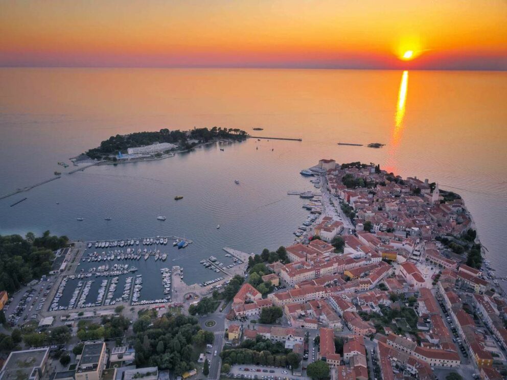 aerial view of the coastal town of Poreč, Croatia, at sunset. The image shows the historic old town on a peninsula, a marina filled with boats, and a small island in the bay. The setting sun casts a golden glow over the Adriatic Sea and the town's red-tiled roofs.