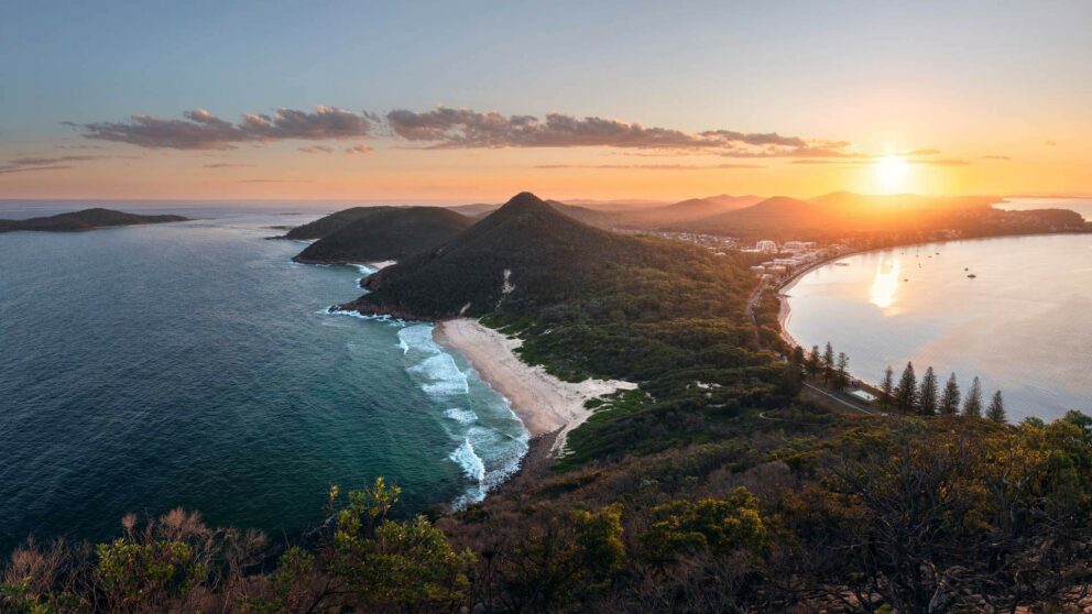 A breathtaking sunset view over Port Stephens, Australia, showing a long stretch of coastline, forested hills, and a golden sun setting over the calm bay waters.