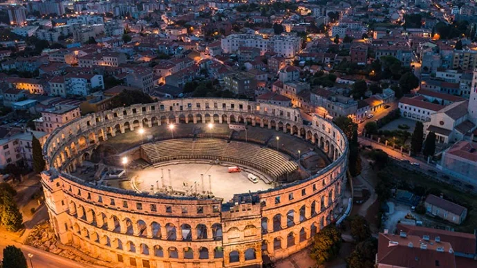 Aerial view of the Pula Arena, a well-preserved Roman amphitheater, at dusk. The ancient structure is illuminated, standing out against the surrounding city lights of Pula, Croatia.