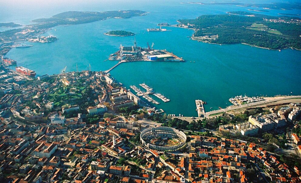 An aerial view of the coastal city of Pula, Croatia. The image shows the city's red-roofed buildings surrounding a well-preserved Roman amphitheater, with a large harbor and a shipyard in the background.