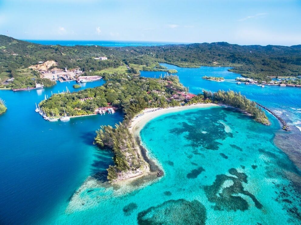 Aerial view of Roatán Island, Honduras, featuring turquoise coral reefs, lush greenery, and boats anchored in the crystal-clear Caribbean waters.