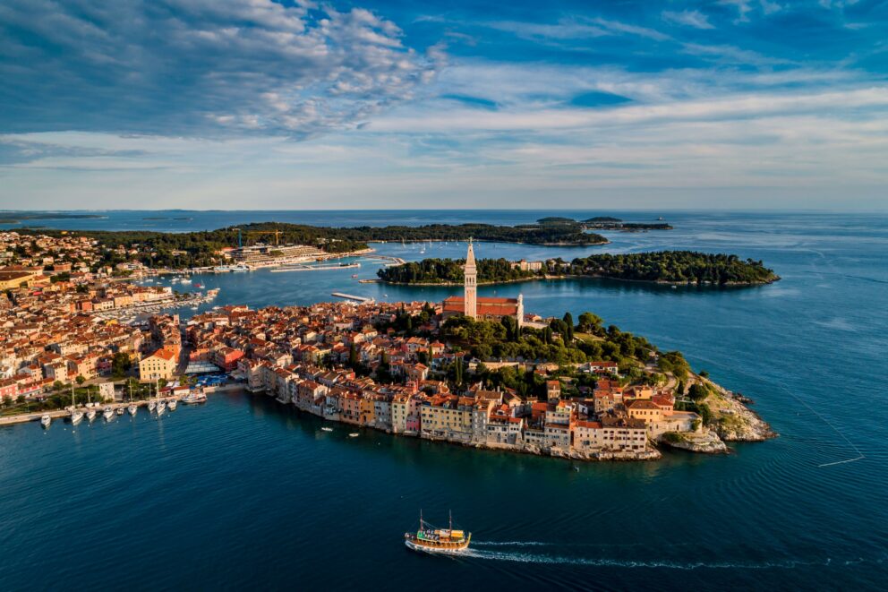 Aerial view of the coastal town of Rovinj, Croatia, located on the Istrian peninsula. The old town is situated on a small peninsula that juts into the Adriatic Sea, with colorful buildings tightly clustered together and a prominent church tower, the Church of St. Euphemia, at its highest point. The deep blue sea surrounds the town, with several small islands visible in the background. A small boat can be seen in the foreground, leaving a wake behind it. The sky is blue with some scattered white clouds.