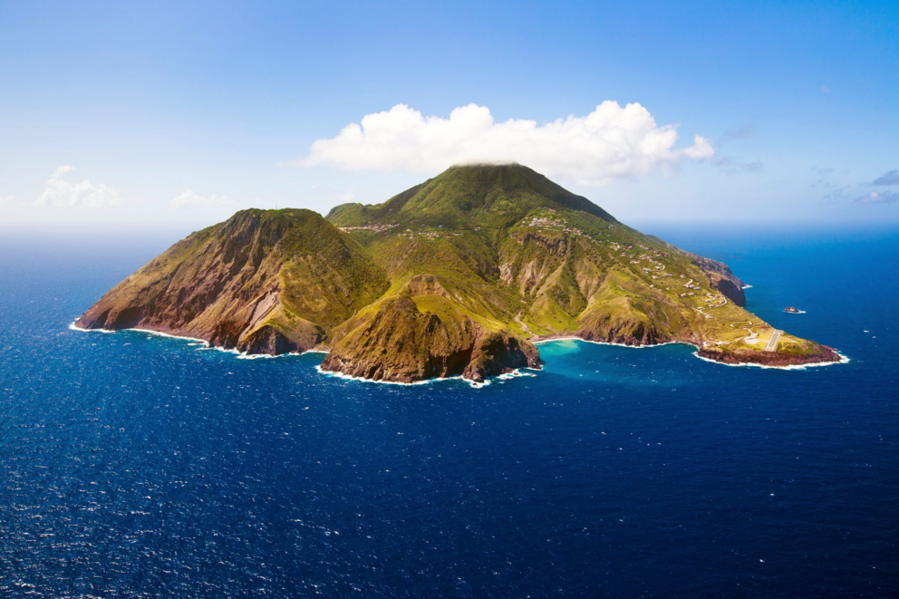 Aerial view of a lush green volcanic island, likely in the Caribbean, surrounded by turquoise ocean under a partly cloudy sky