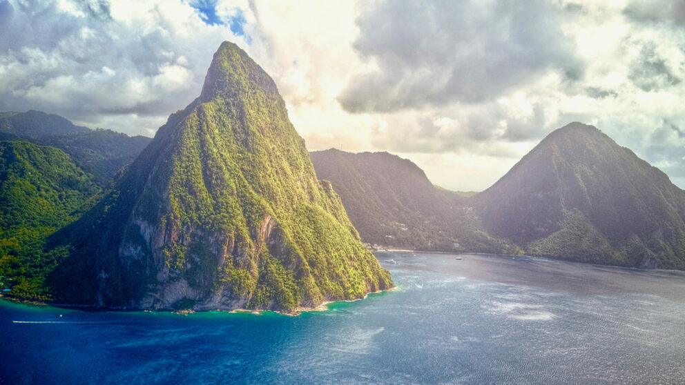 A scenic view of the Pitons, Gros Piton and Petit Piton, in St. Lucia, rising dramatically from the blue Caribbean Sea under a bright, partly cloudy sky.