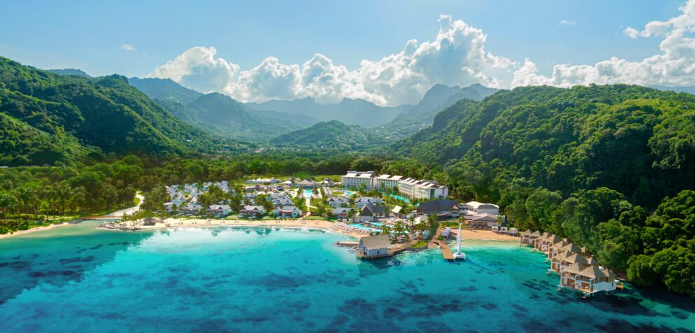Aerial view of a luxury beach resort with overwater bungalows and lush green mountains in the background.