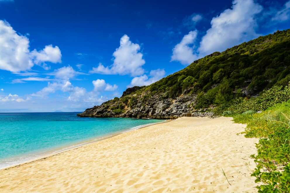 A bright, sunny beach with turquoise water, golden sand, and a lush green hill on the right.