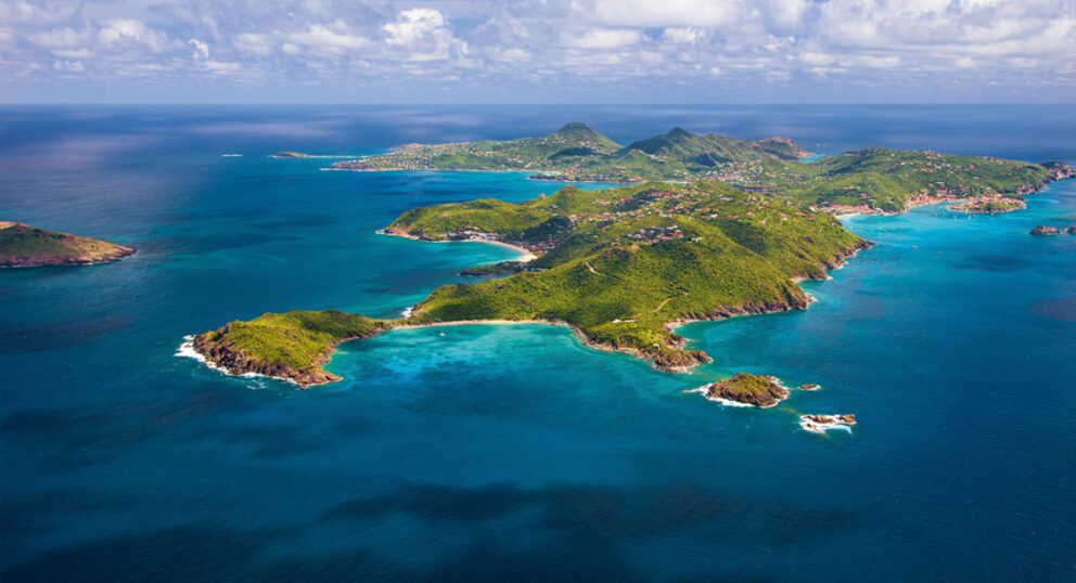 Aerial view of a lush, green, hilly Caribbean island surrounded by clear turquoise and deep blue ocean water.
