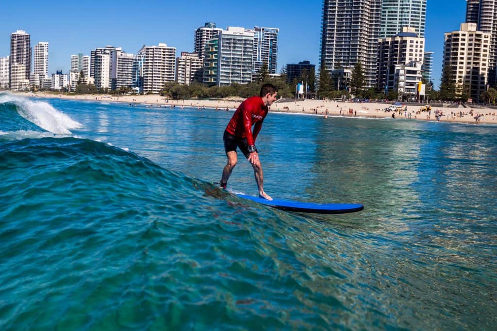 A surfer riding a gentle wave on a blue surfboard near Surfers Paradise Beach, with tall city buildings and a sandy shoreline full of beachgoers in the background under a clear blue sky.