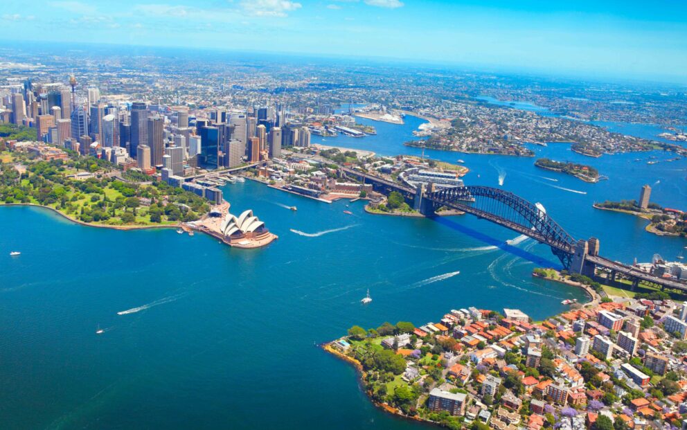 Aerial view of Sydney Harbour showing the Sydney Opera House, the Sydney Harbour Bridge, and the surrounding cityscape with boats moving through the deep blue water under a clear sky.