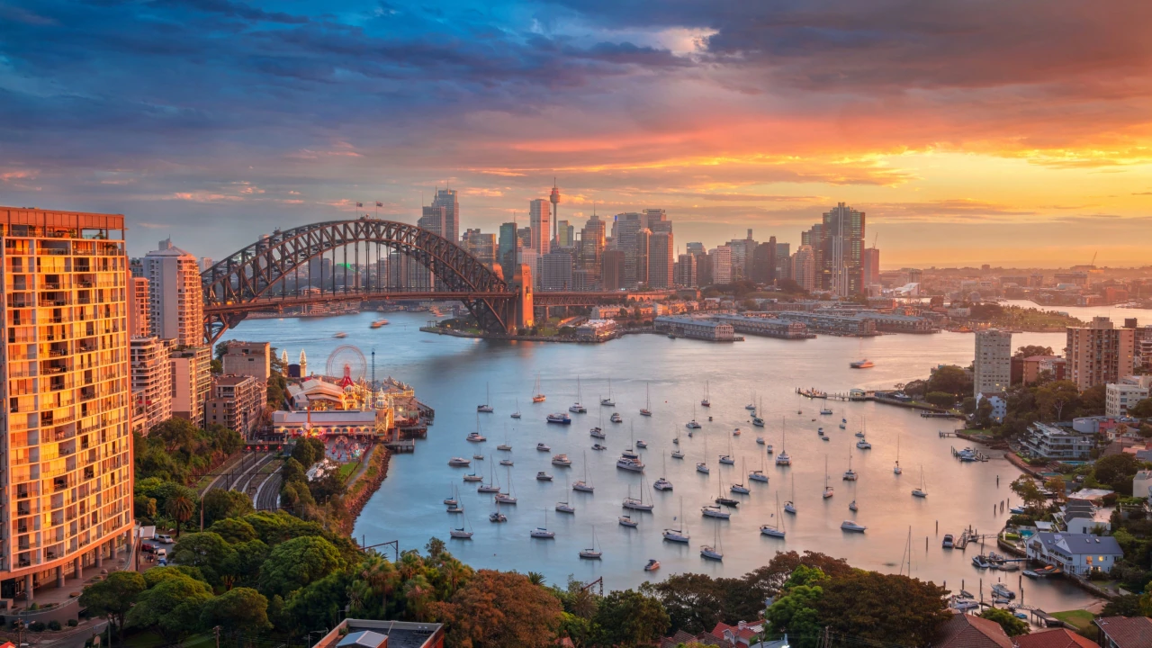 A stunning sunset view over Sydney Harbour, Australia, showcasing the iconic Sydney Harbour Bridge, city skyline, and boats anchored in the harbor, with warm golden and pink hues reflecting on the water.